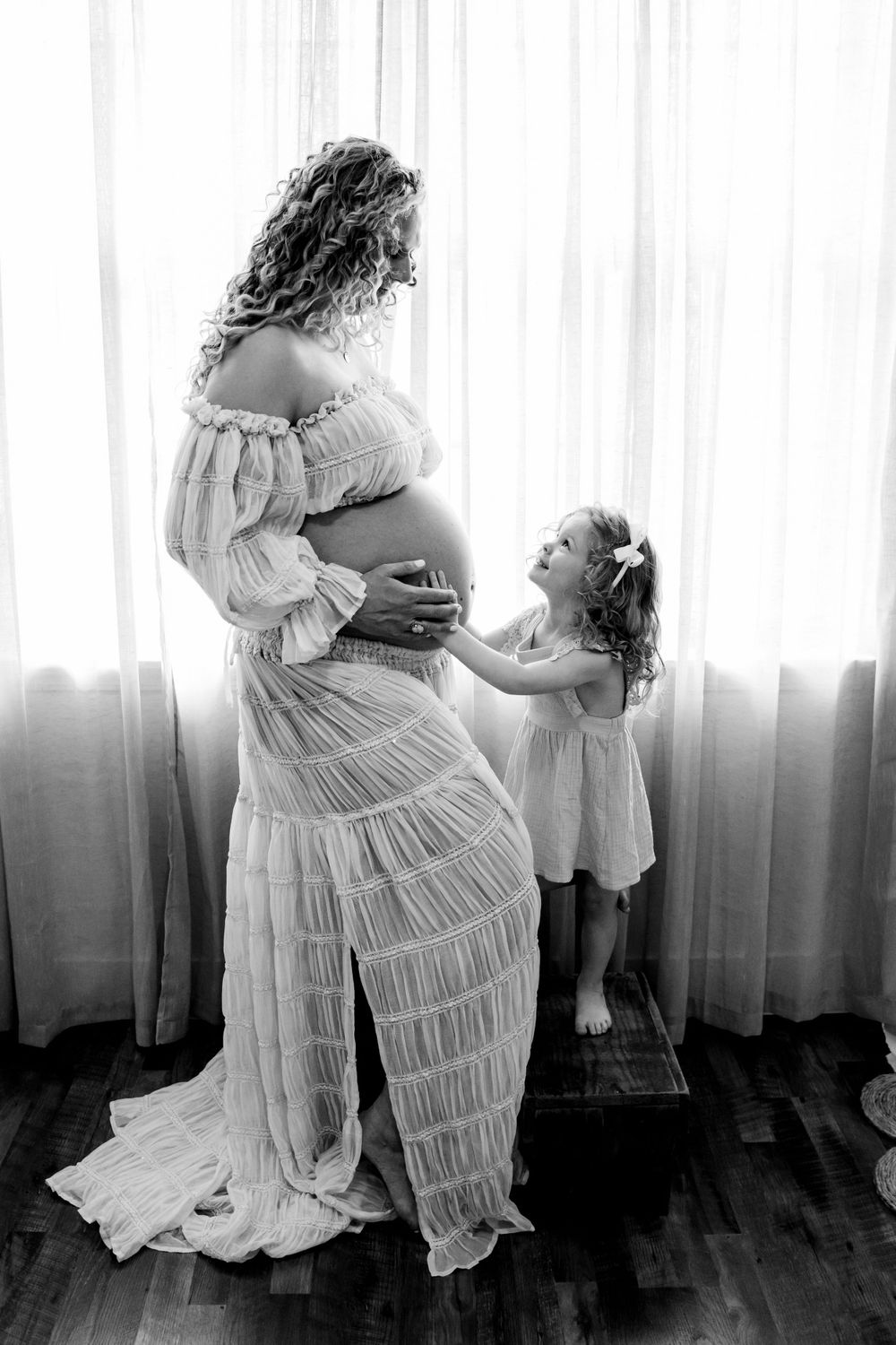 Black and white maternity photo with child touching mother's pregnant belly by window.