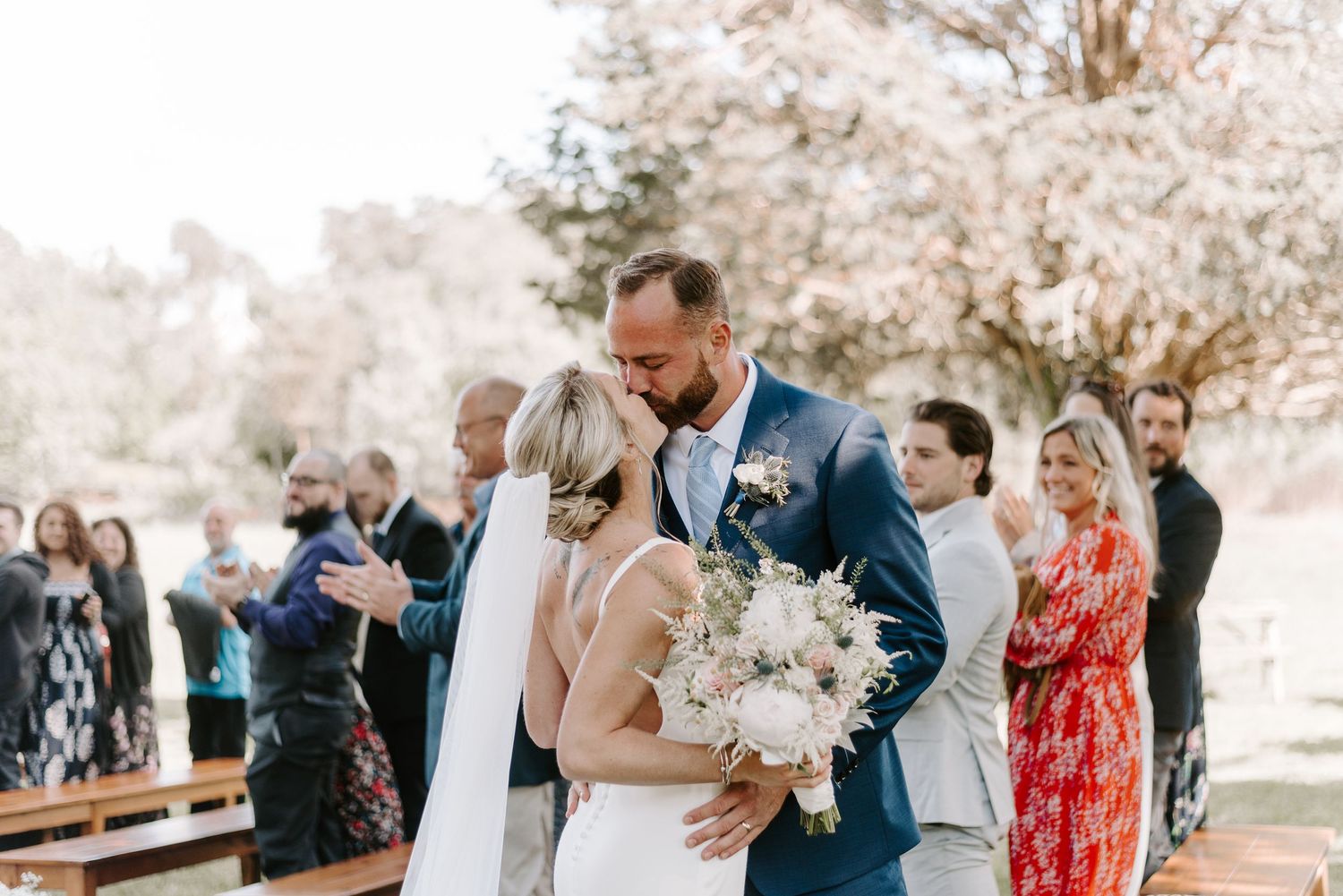 Wedding party gathers under blooming cherry blossom trees in a garden setting.