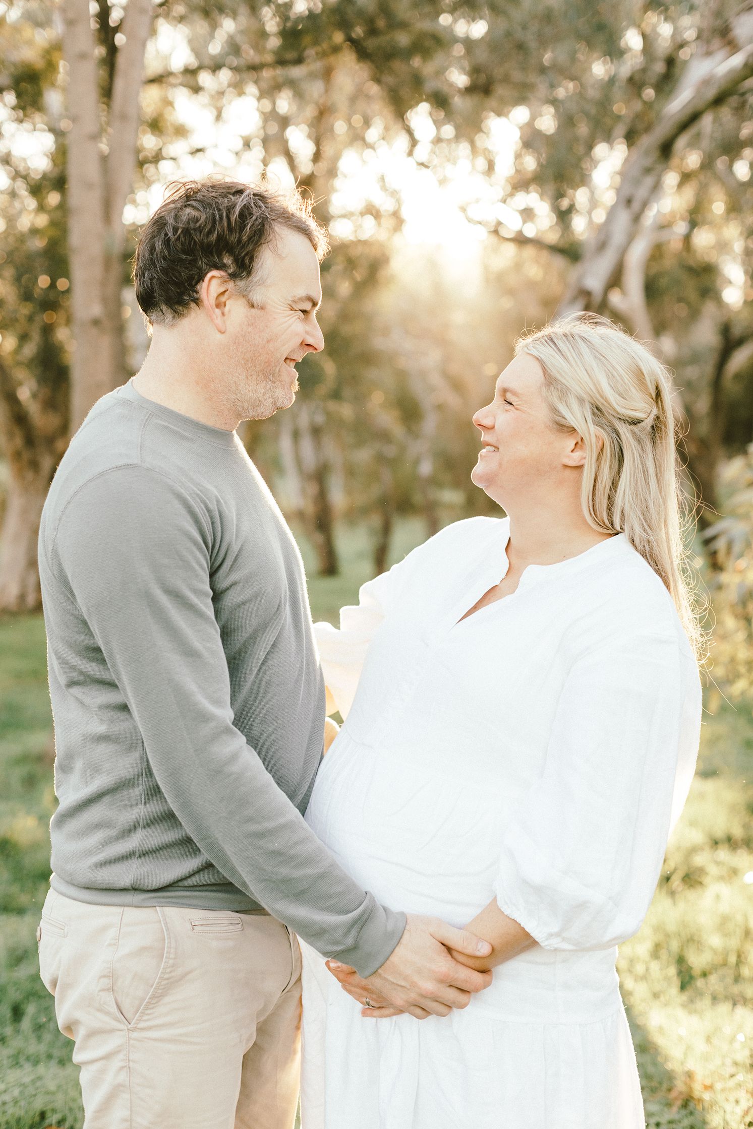 A sweet maternity photoshoot in a sun-drenched field with a couple dressed in grey and white embracing.