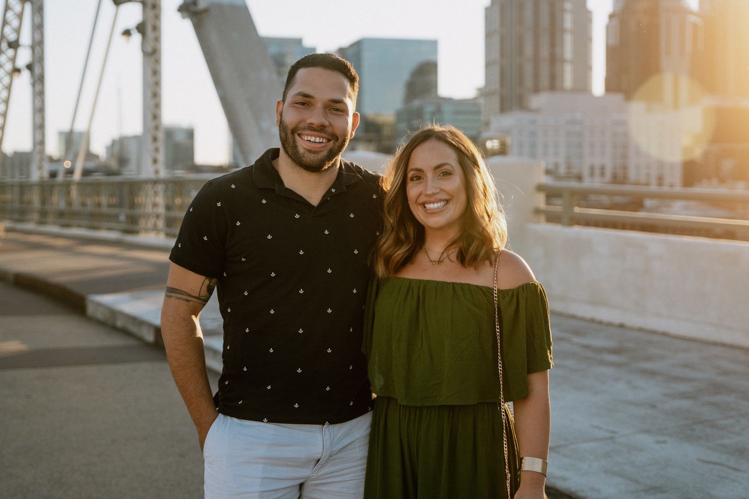 A newly engaged couple smiles together on a rooftop with the city skyline behind them.
