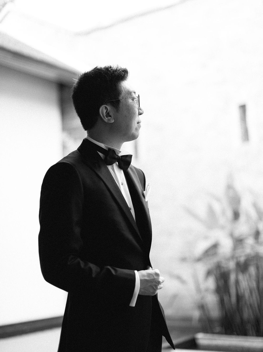 A groom in a tuxedo stands in front of a building in Phuket, Thailand, eagerly awaiting his special day.