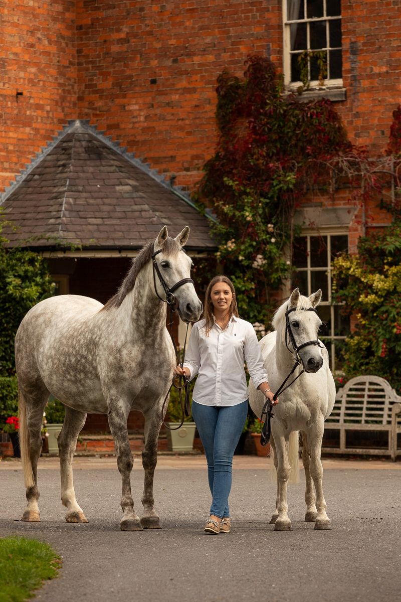 Two grey horses and their handler stand in front of a brick country house with climbing vines.