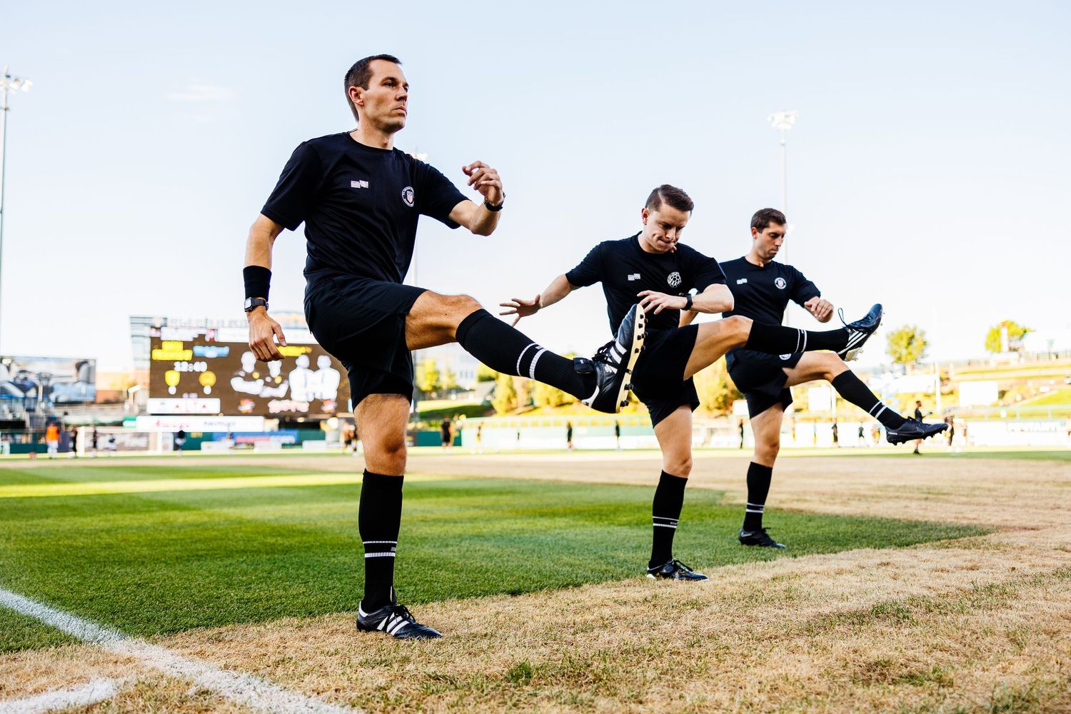 Referees performing synchronized high kicks on a soccer field during warmup.