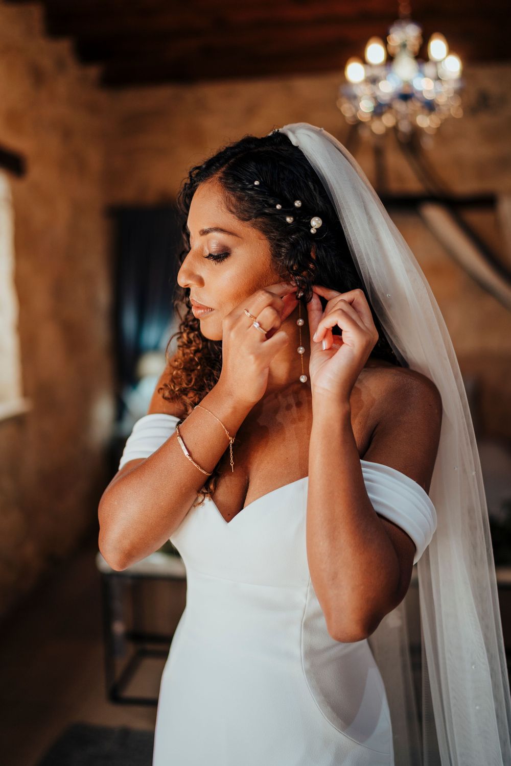 Bride in an off-shoulder white dress puts on earrings while getting ready under a crystal chandelier.