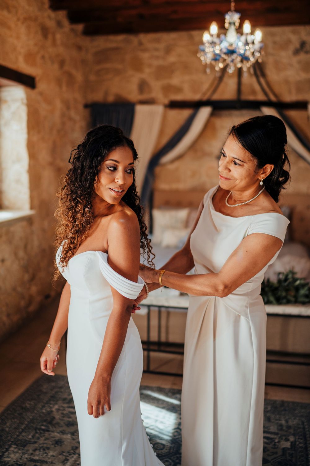 Two women in elegant white dresses share an intimate moment during wedding preparations in a rustic stone venue.