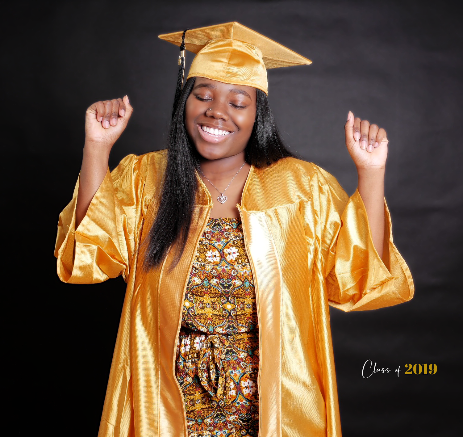 Graduate in gold cap and gown celebrates with outstretched arms against dark background.