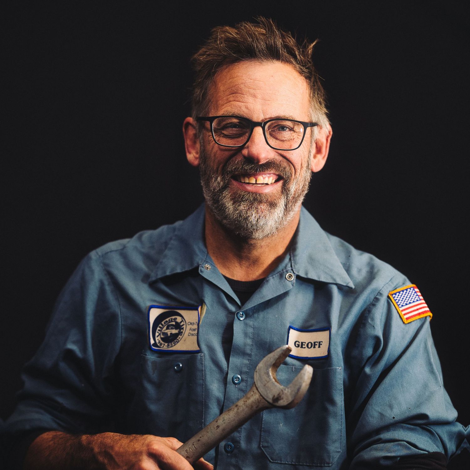 Smiling maintenance worker in blue uniform and glasses poses against dark background.