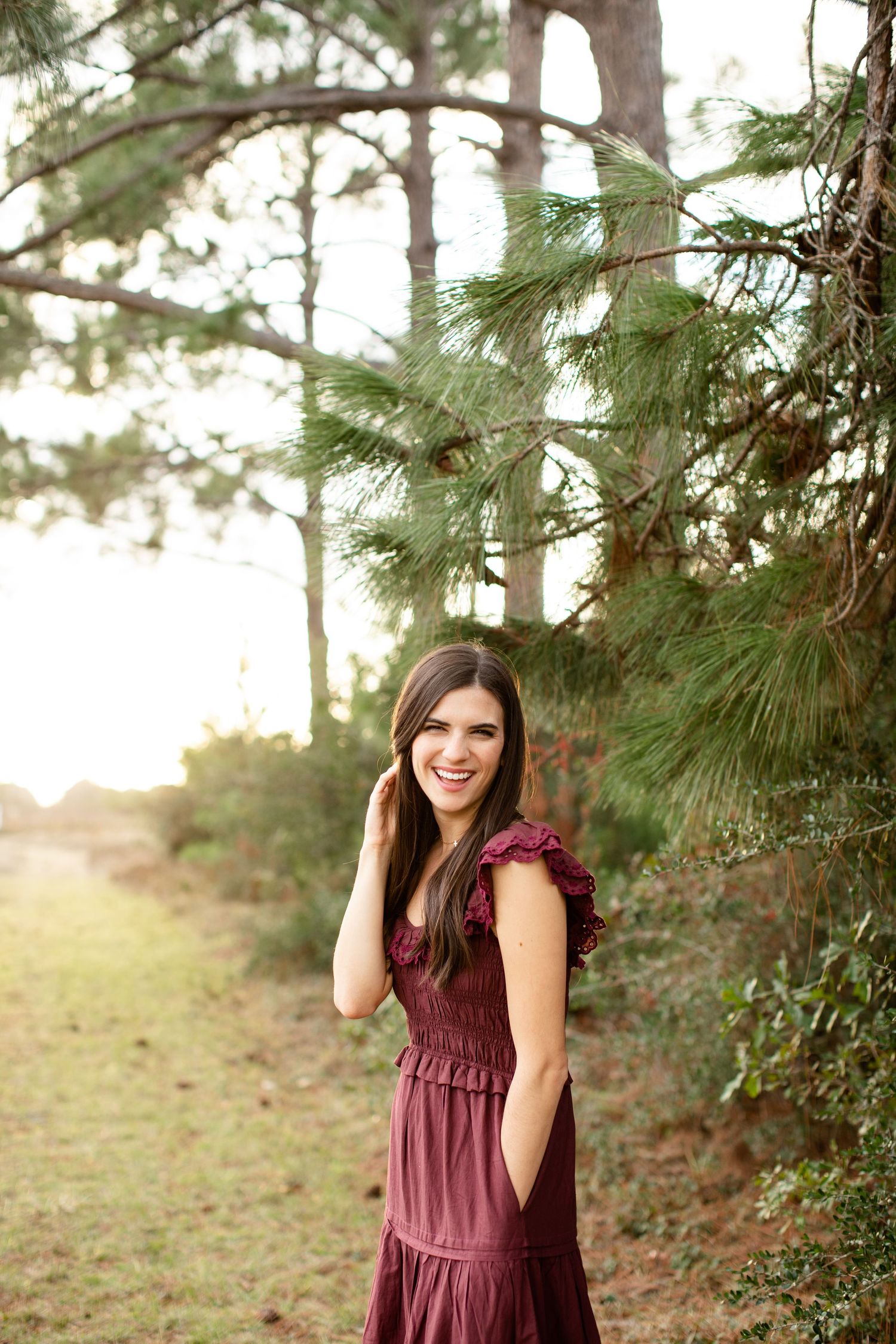 Woman in red dress stands in golden sunlight beneath pine trees.