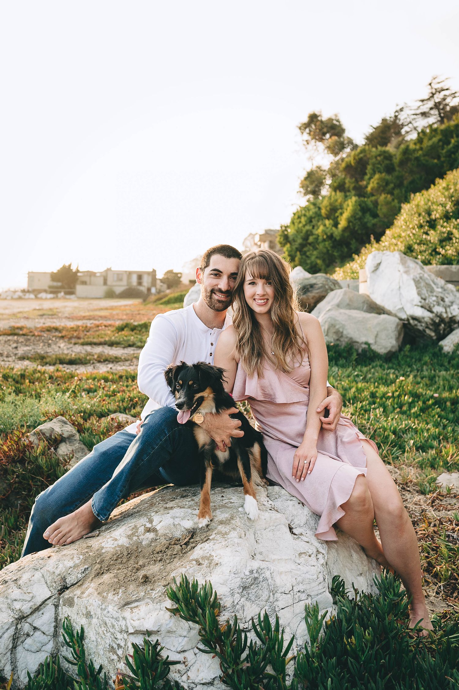 Couple with small black dog poses on rocky outcrop by coastal landscape.