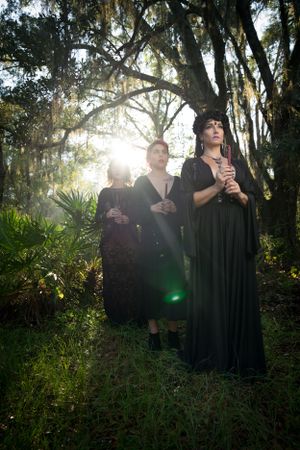 Figures in dark clothing pose atmospherically in a misty forest setting with dramatic backlighting.