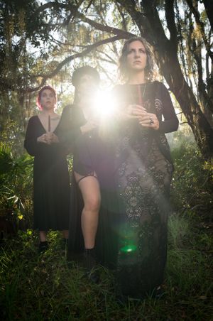 Sunlight streams through trees creating ethereal lighting effects around dark-clothed figures in nature.