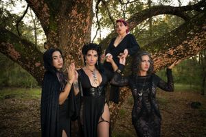 A group in dark gothic attire poses beneath a large twisted tree in an autumn forest setting.