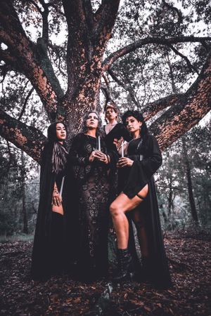 Dramatic forest photoshoot with figures in flowing black clothing posed around a gnarly tree trunk.