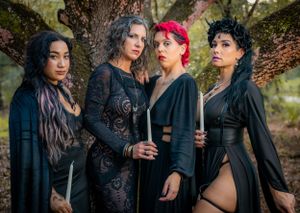 Gothic-styled group holding candles while posing beside a large tree in dark clothing.