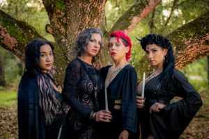 People in black gothic clothing pose dramatically around a tree in a mystical forest scene.