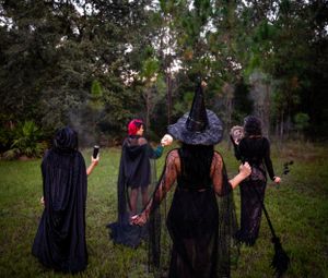 Figures in dark cloaks and witch hats gather mysteriously in a wooded outdoor setting at dusk.