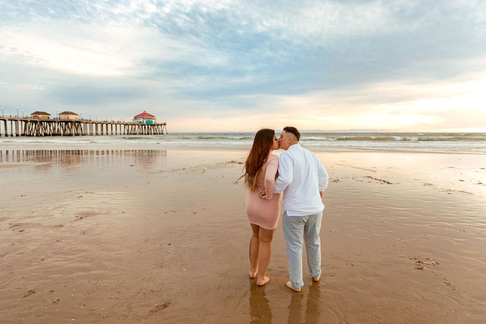 A romantic couple stands on a sandy beach at sunset with a wooden pier stretching into the ocean in the distance.