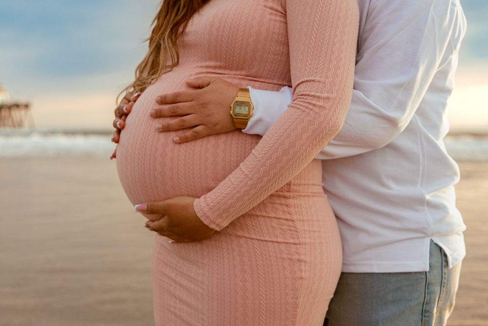 A maternity photo showing a pregnant woman in pink sweater embracing her belly with partner standing on beach at sunset.