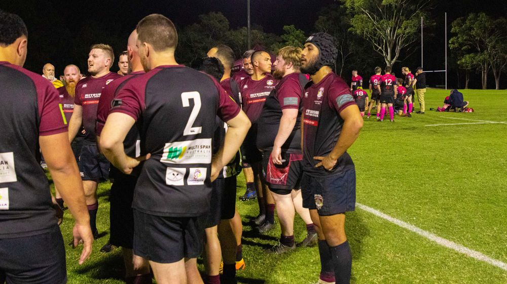 Rugby players in maroon uniforms huddle on the field during a nighttime game.