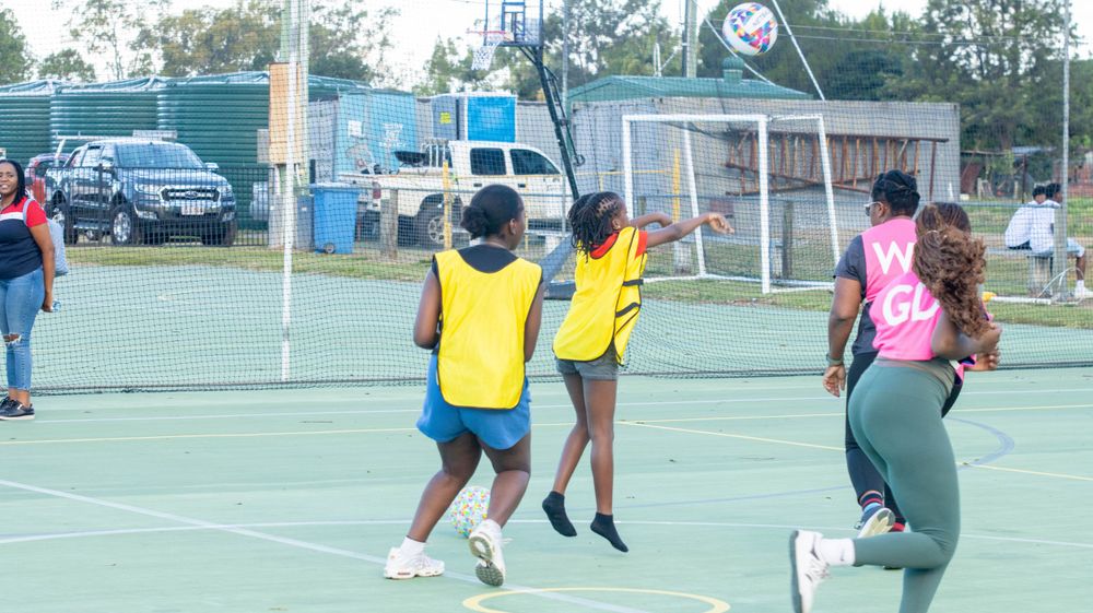 Players in yellow and pink jerseys compete during a netball game with a goal post visible.