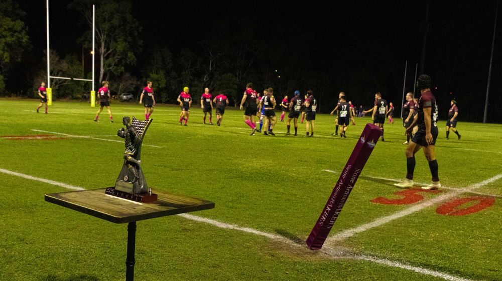 Two trophies displayed on stands with rugby players visible in the background on a lit field.
