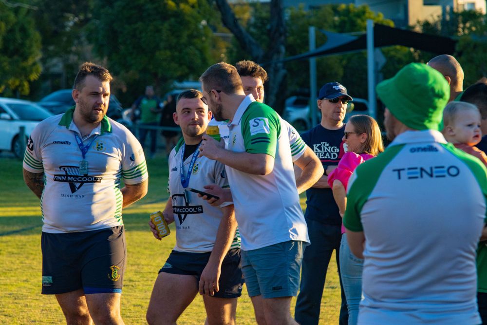 Athletes in sports uniforms gather and socialize on a grass field at sunset.