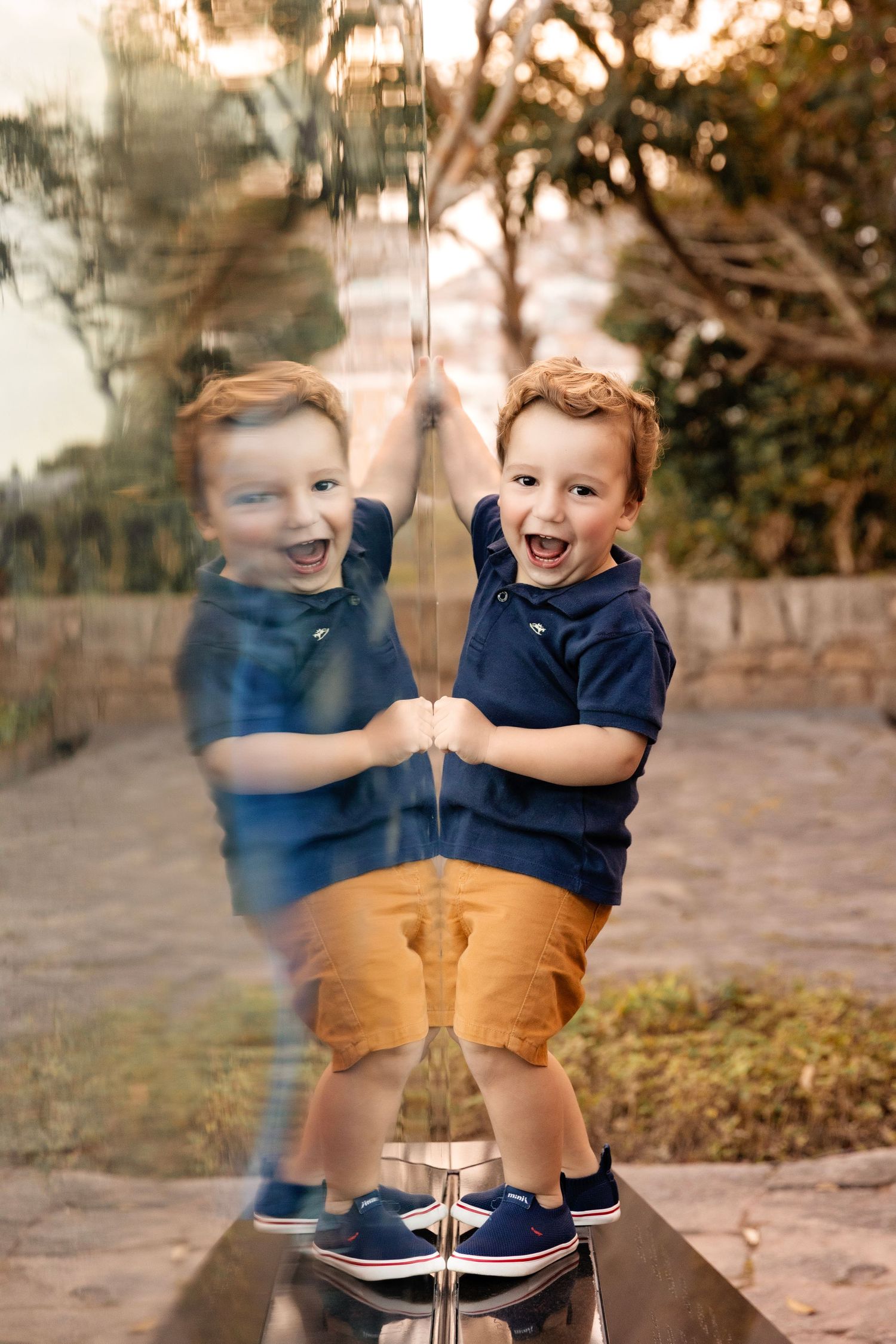 A creative mirror reflection photo of a child laughing in navy blue shirt and khaki shorts.