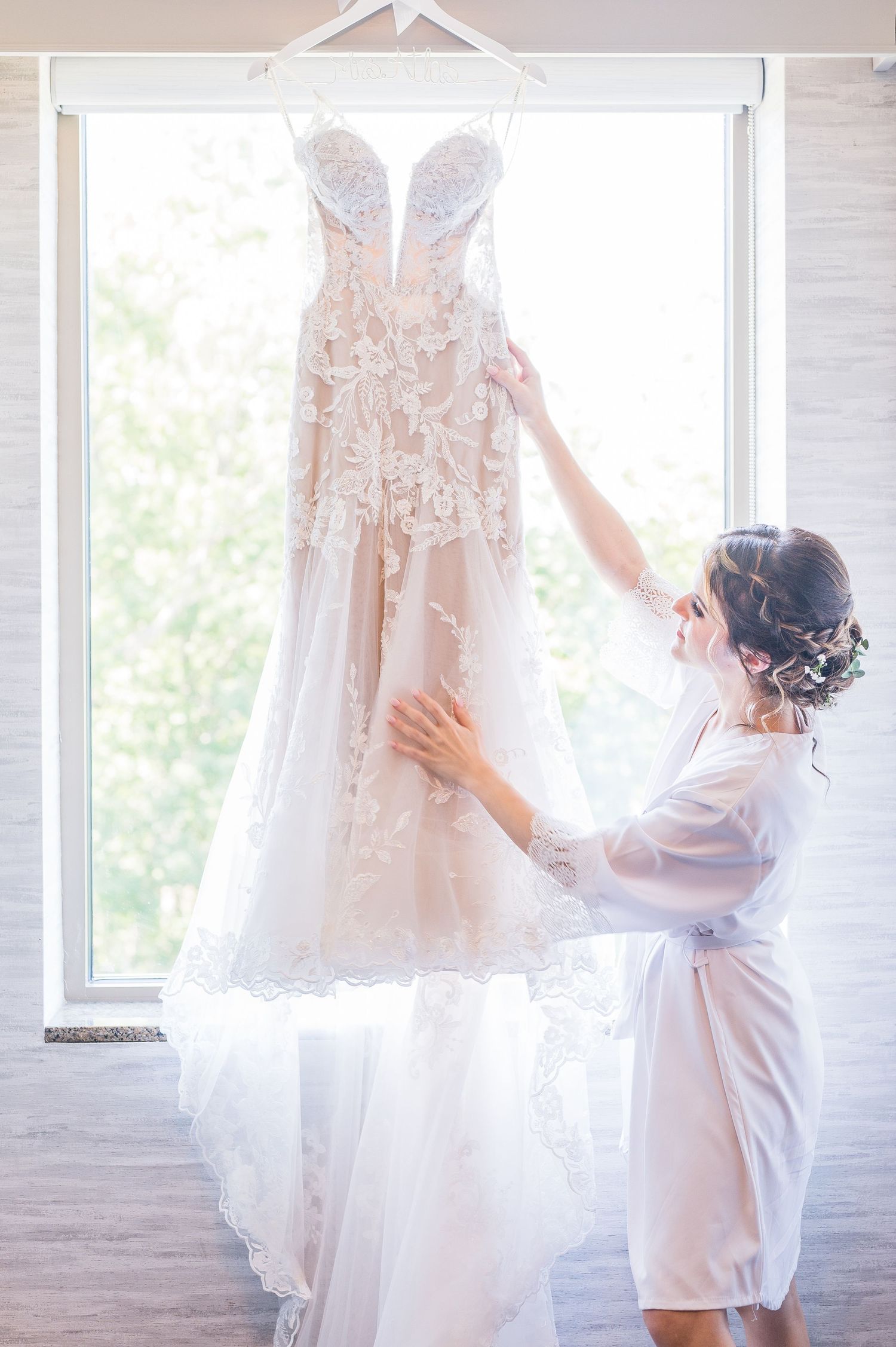 Bride in her robe admiring her wedding dress hanging from the window.