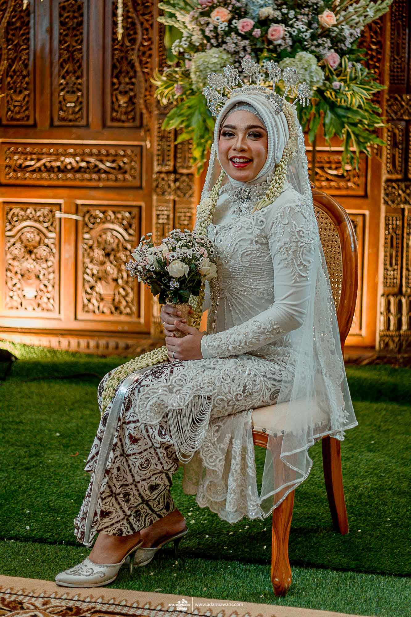 Muslim bride in ornate white lace dress and hijab poses with bouquet against carved wooden backdrop.
