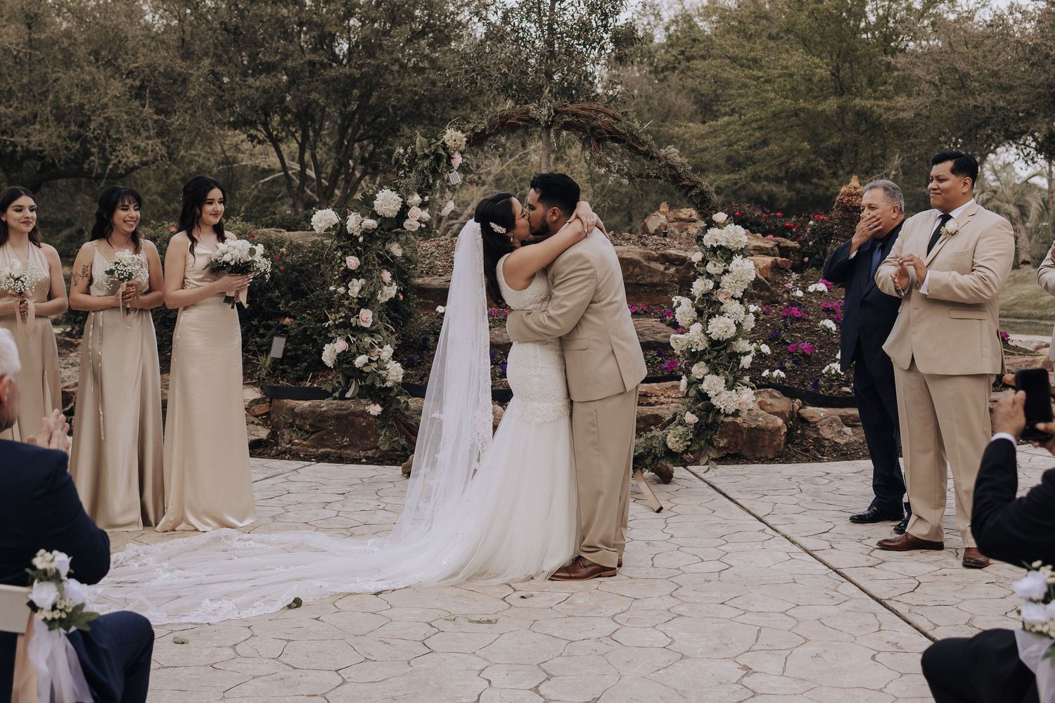 Wedding ceremony participants gather around a floral arch during an outdoor celebration.