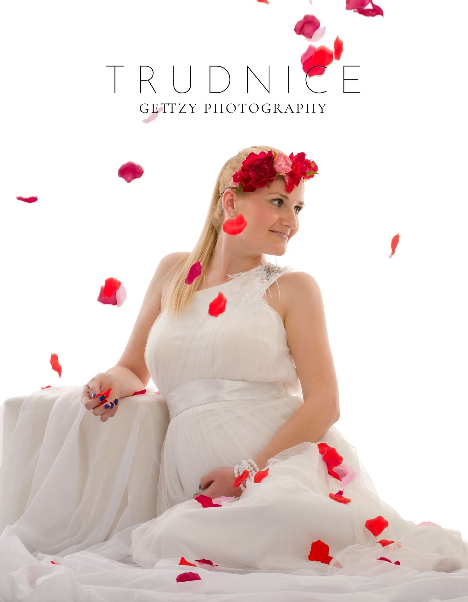 A romantic maternity photo with red rose petals falling around a woman in a white dress.