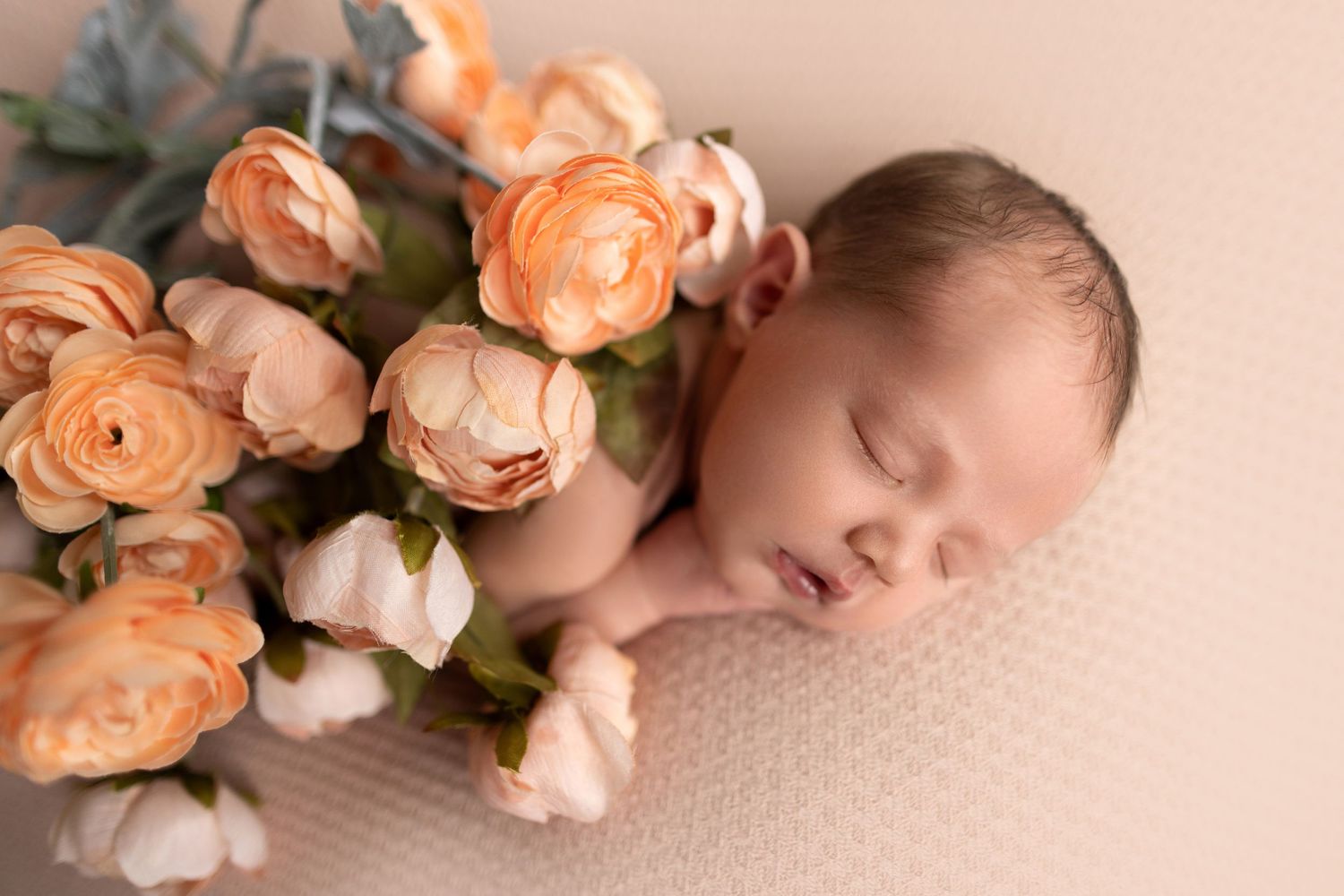 Sleeping newborn baby nestled among peach-colored roses on soft pink fabric.