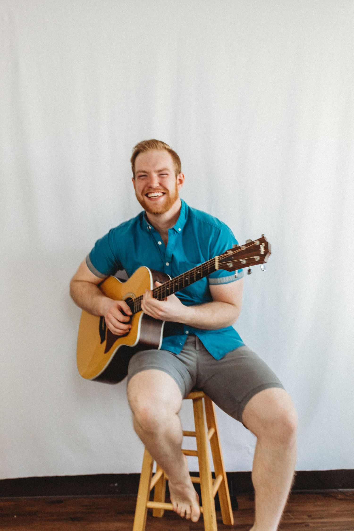 Person in teal polo shirt plays acoustic guitar while sitting on wooden stool.