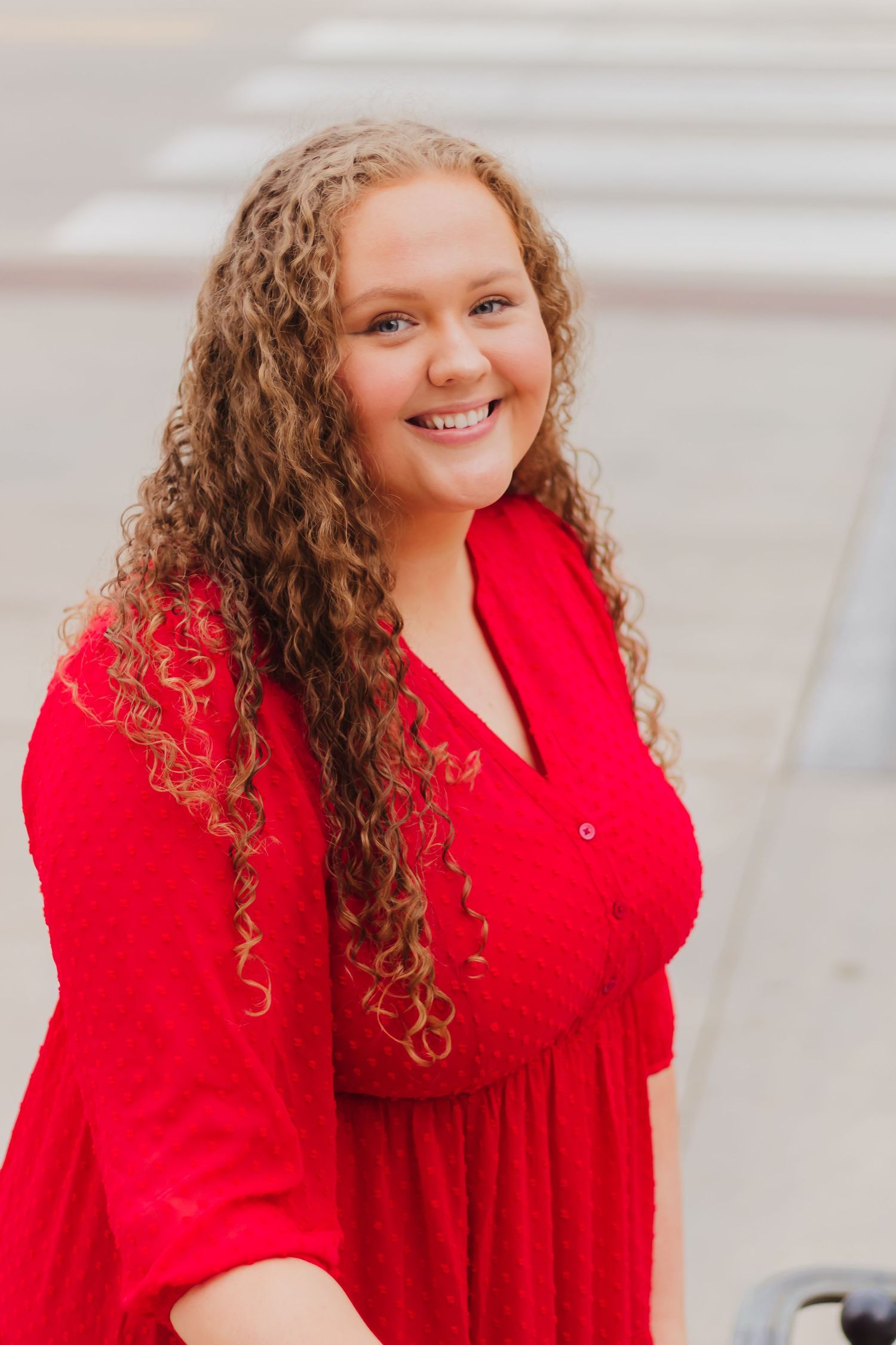Portrait of person in bright red dress with curly hair against light architectural background.