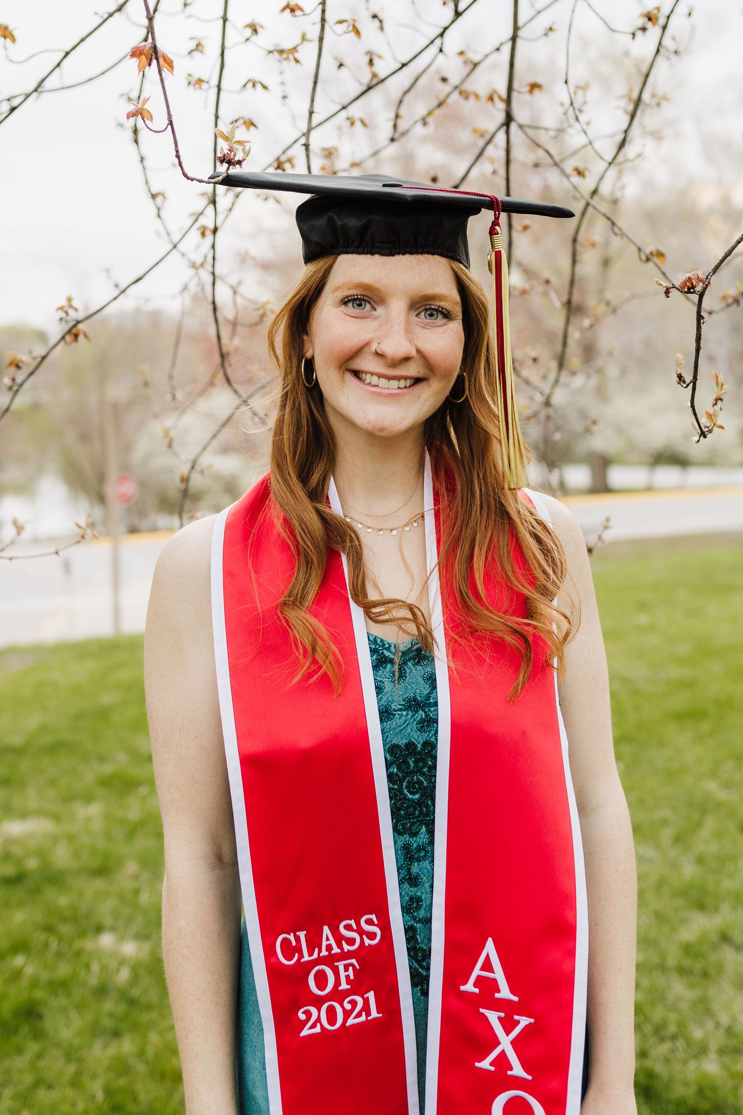 Class of 2021 graduate wearing red stole and cap poses outdoors during spring graduation.