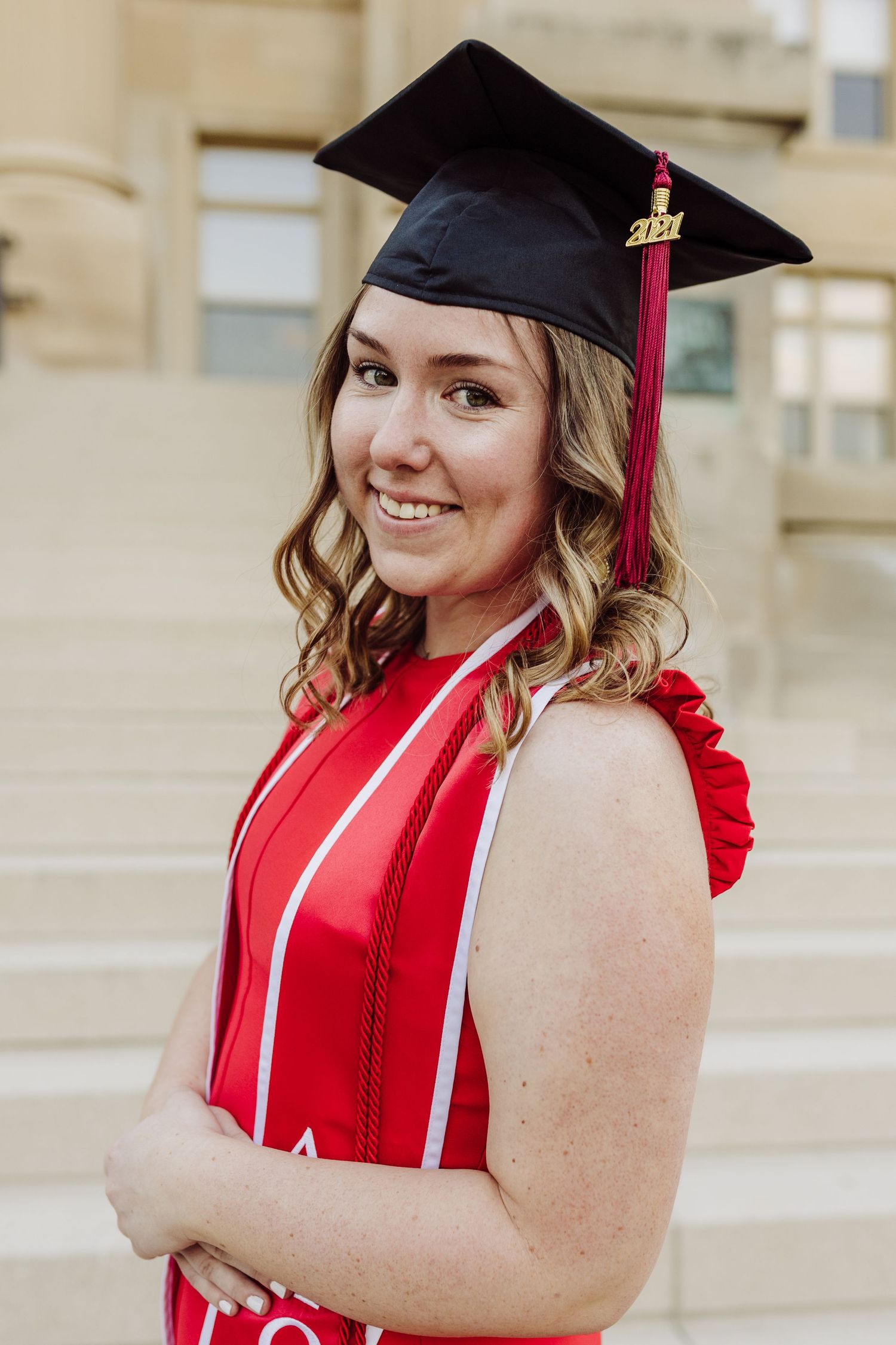 Graduate in red dress and black cap poses on steps during commencement ceremony.