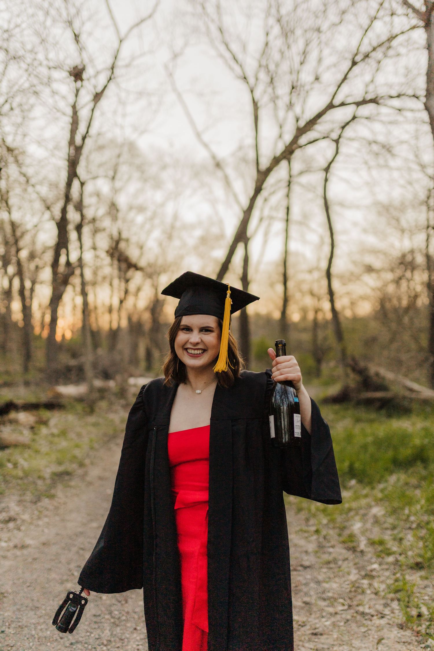 Graduate in red dress and black cap and gown holds diploma while posing on woodland path at sunset.