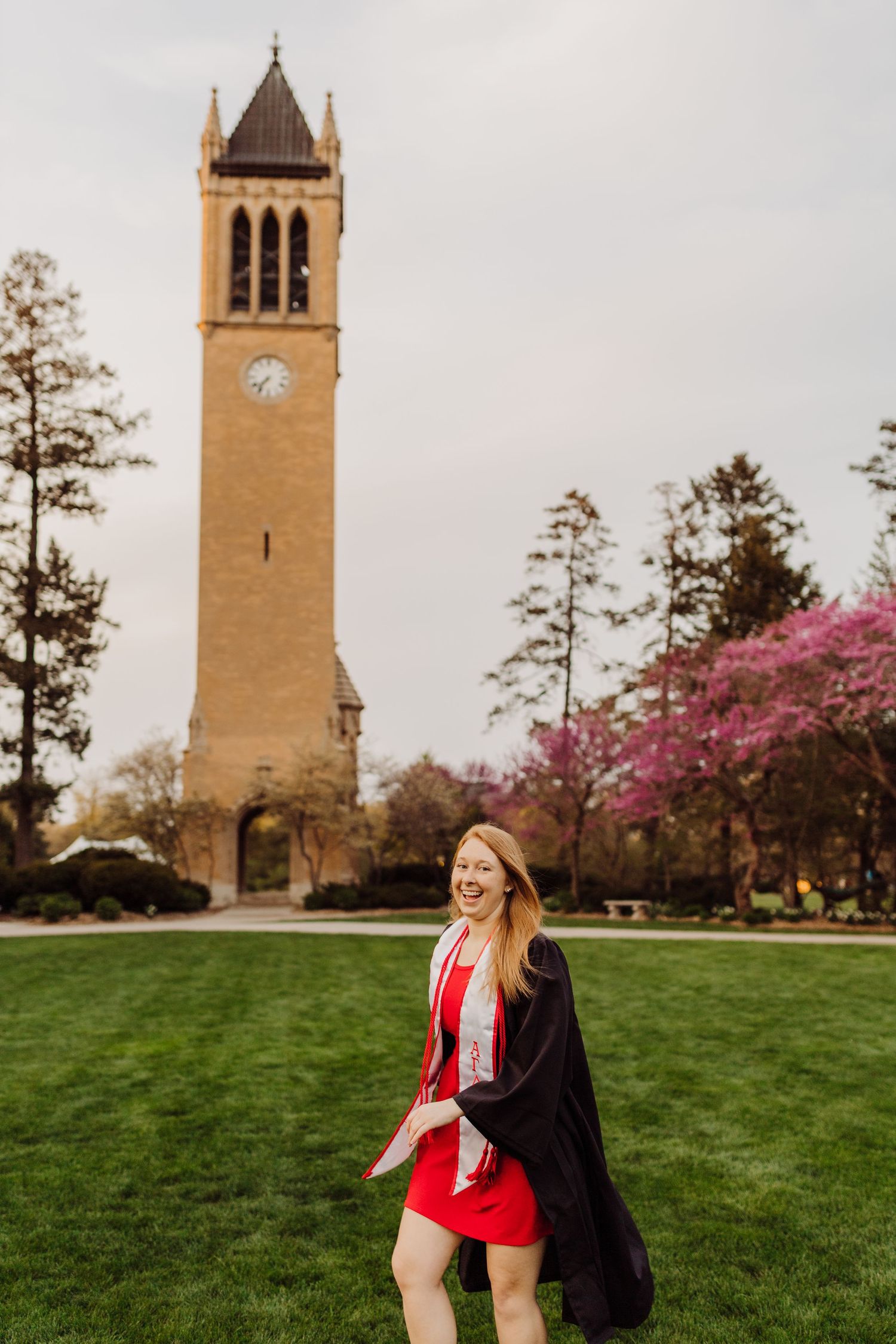 Graduate wearing red dress and black gown poses in front of iconic campus clock tower during spring graduation.