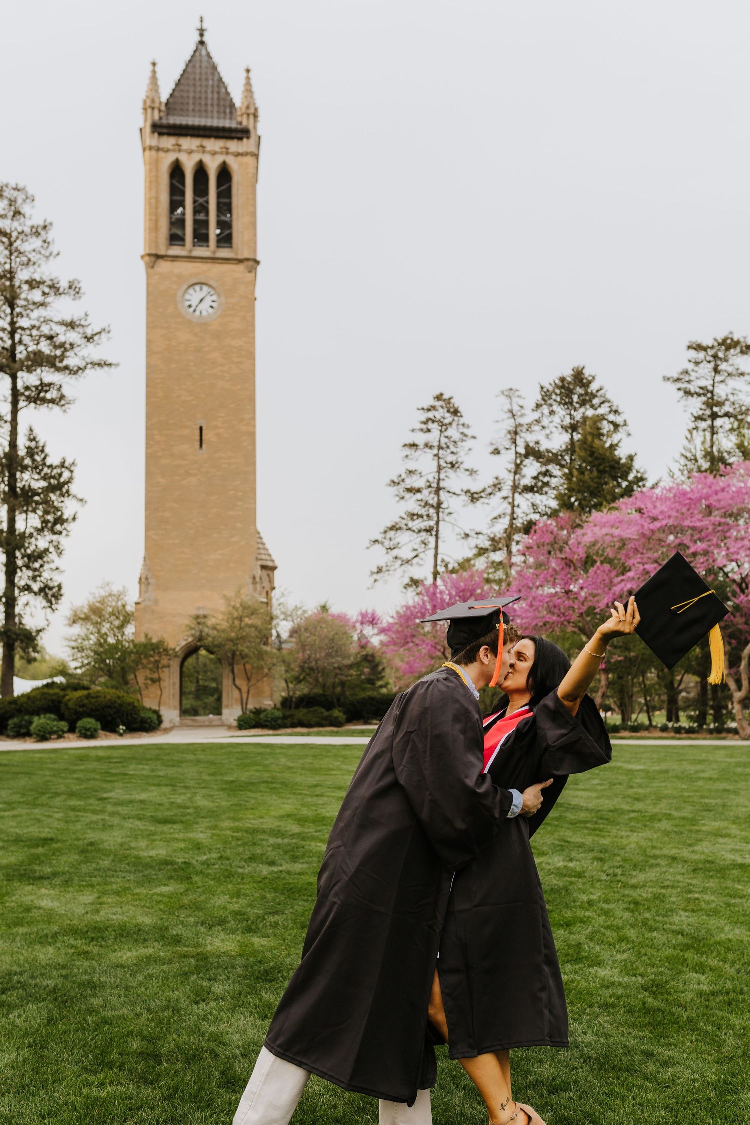 Two graduates celebrate and jump joyfully in front of a historic university bell tower.