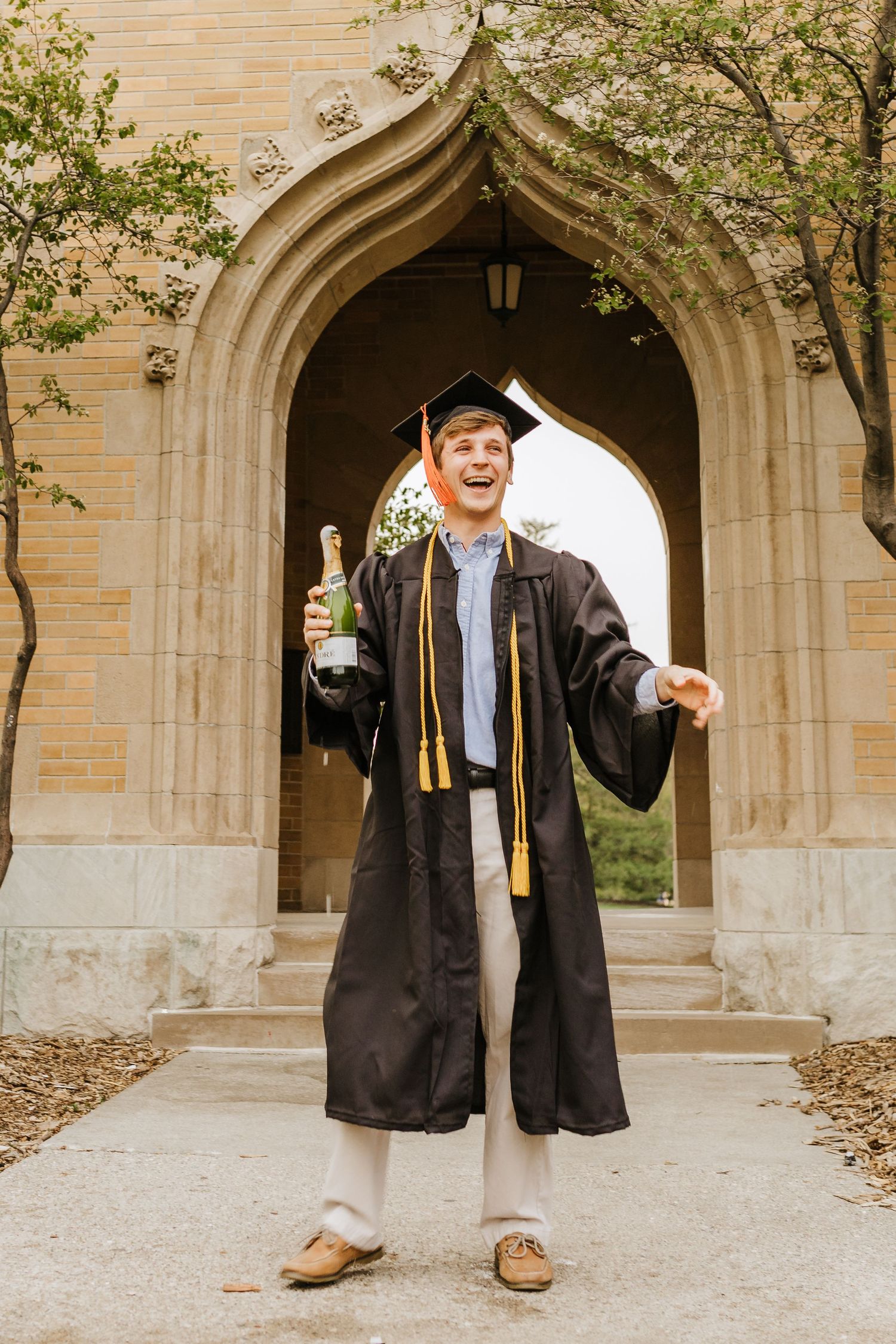 Graduate celebrates under a Gothic archway wearing traditional cap and gown while holding a beverage.