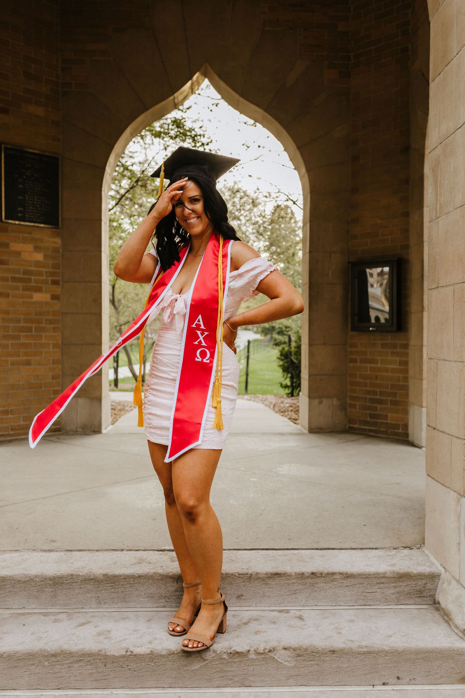 Graduate poses playfully in cap, gown and red sash under a Gothic stone archway.