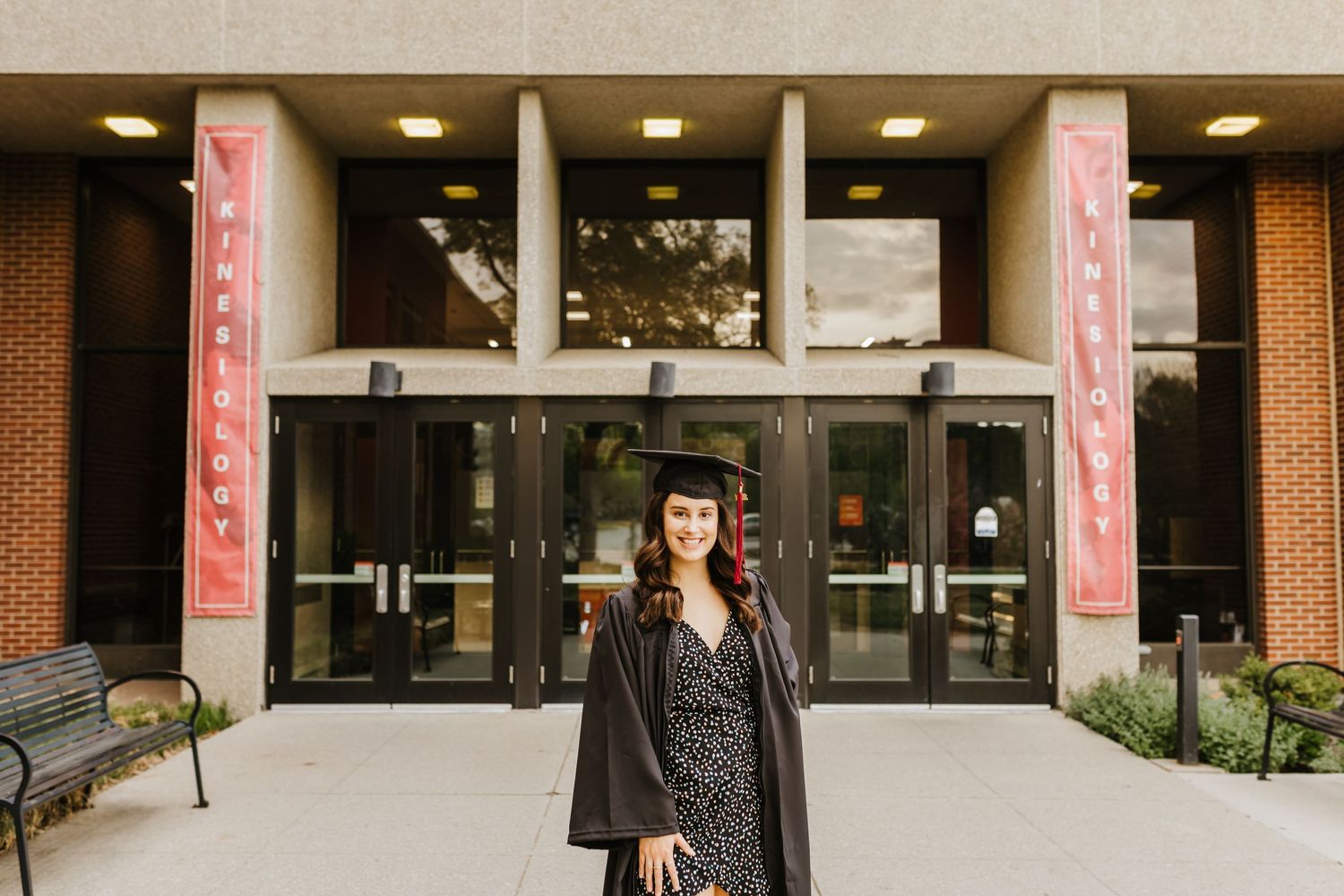 Graduate poses in cap and gown outside a brick university building entrance.