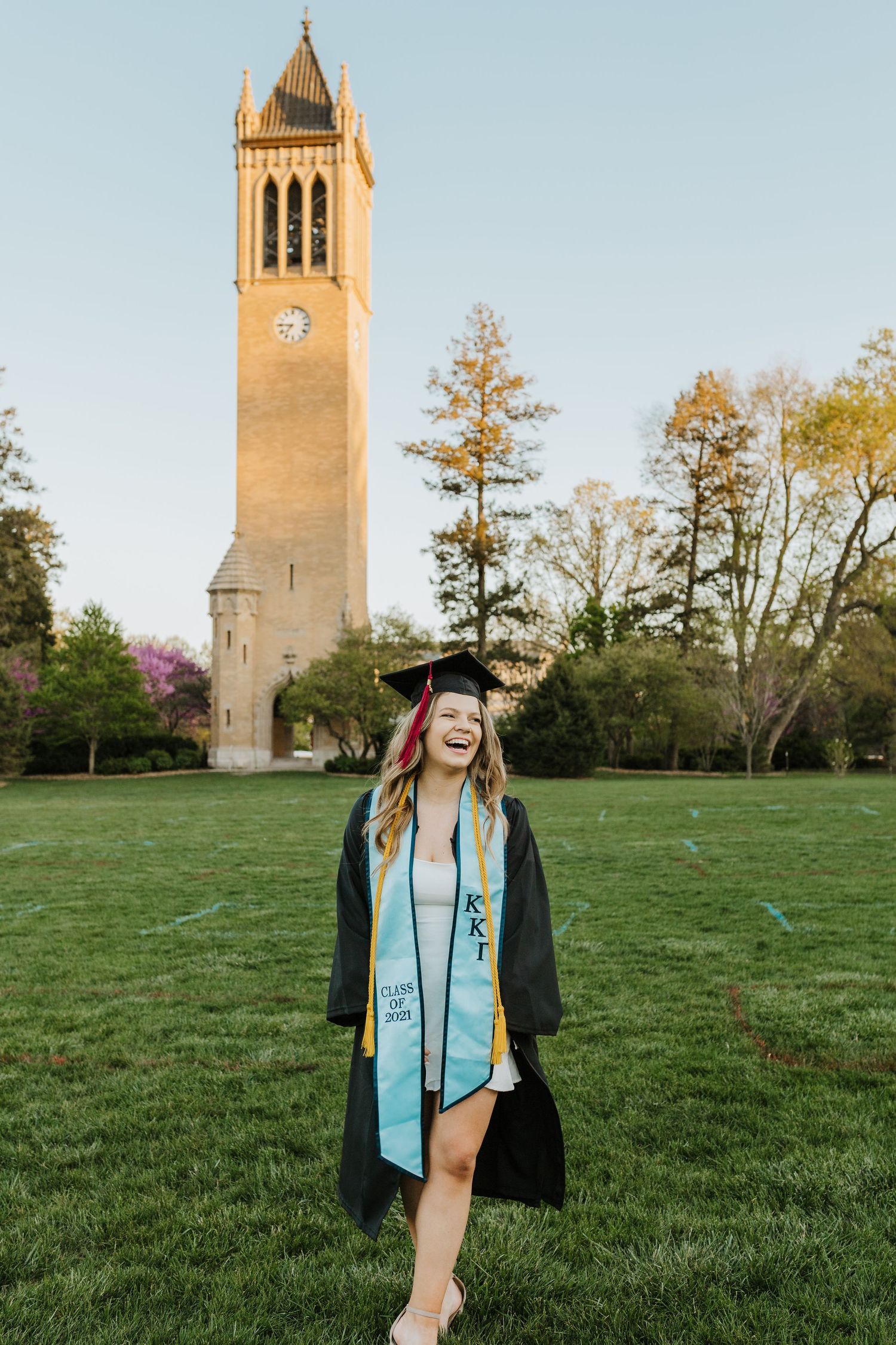 Graduate celebrates in academic regalia in front of a tall brick bell tower during golden hour.