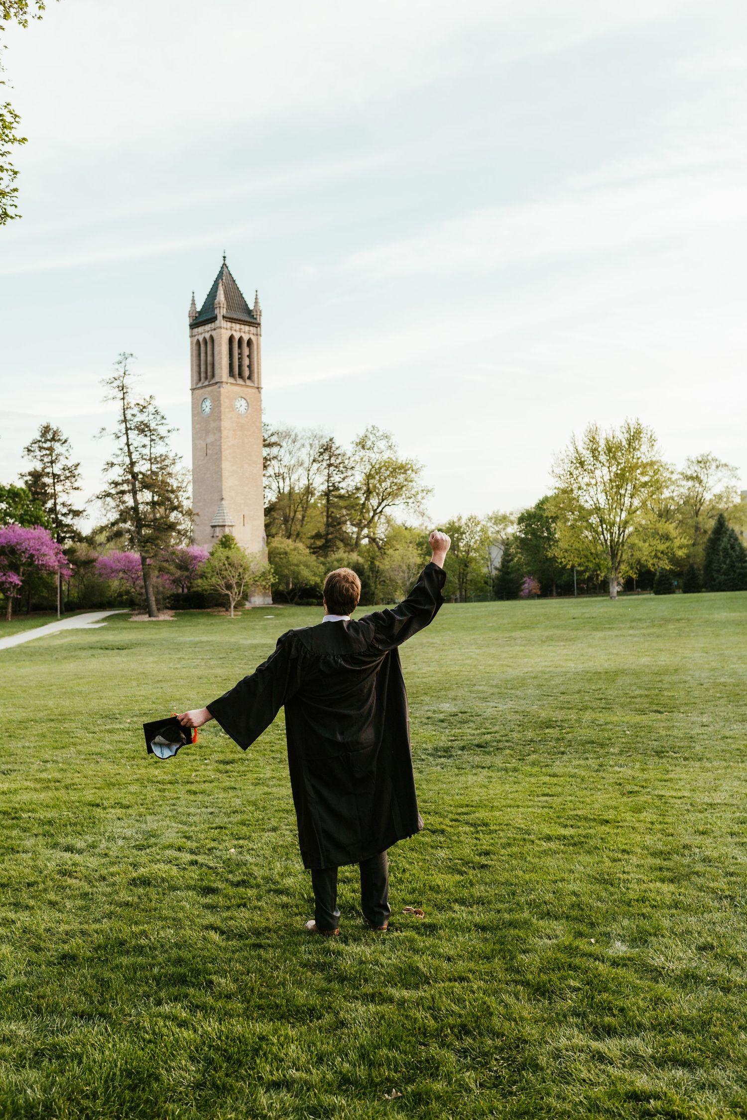 A graduate in black cap and gown celebrates with outstretched arms in front of a tall campus clocktower.