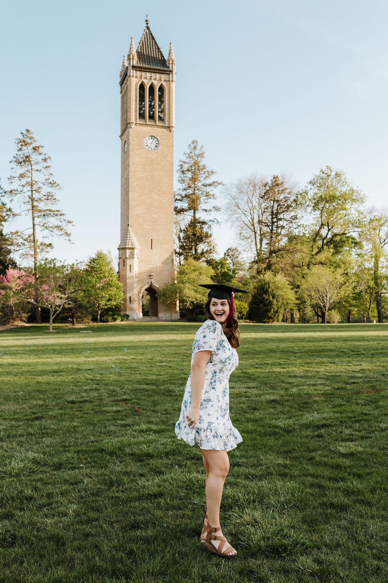 A graduate in a floral dress poses on the lawn in front of the iconic campus clocktower on graduation day.