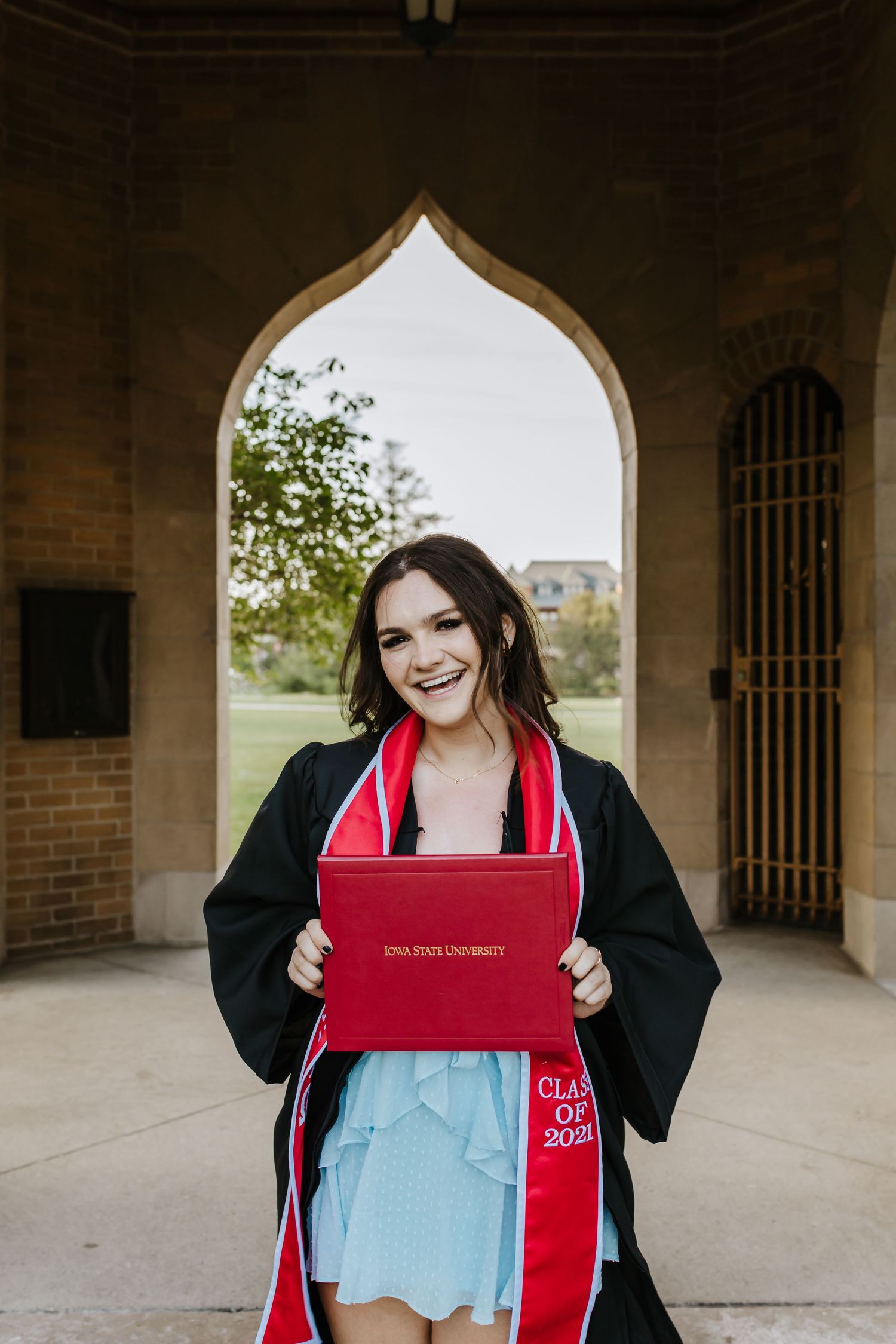 Graduate holding red diploma cover poses under gothic archway in graduation attire.
