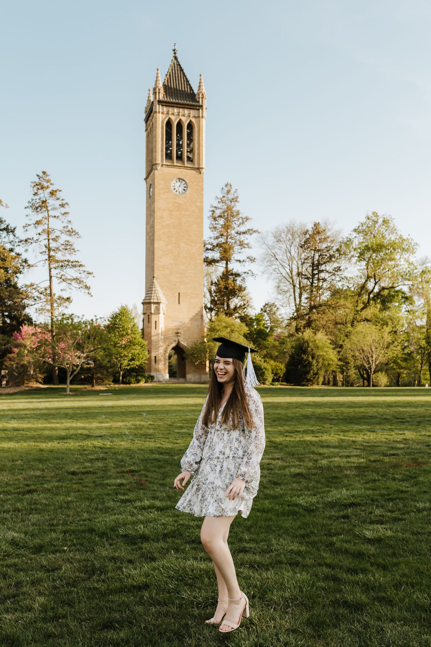 Graduate in floral dress poses with campus clock tower during golden hour.