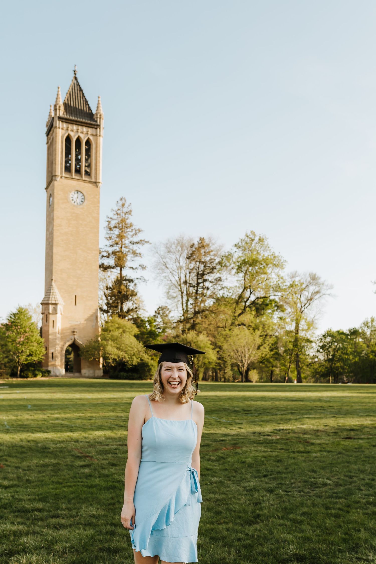 Graduate in light blue dress smiles in front of tall university clock tower on sunny day.
