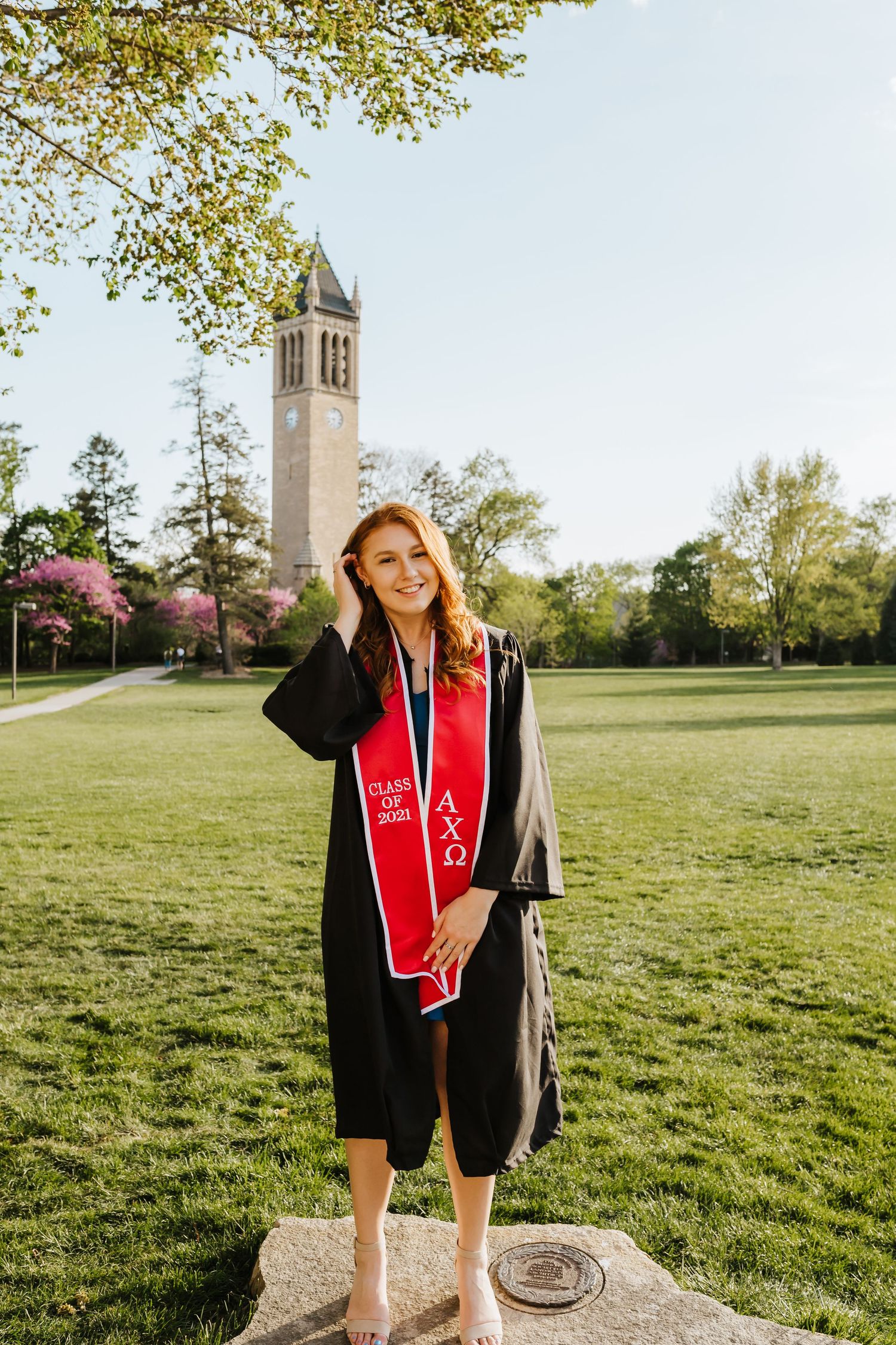 Graduate in traditional black gown and red stole poses near iconic campus clock tower.