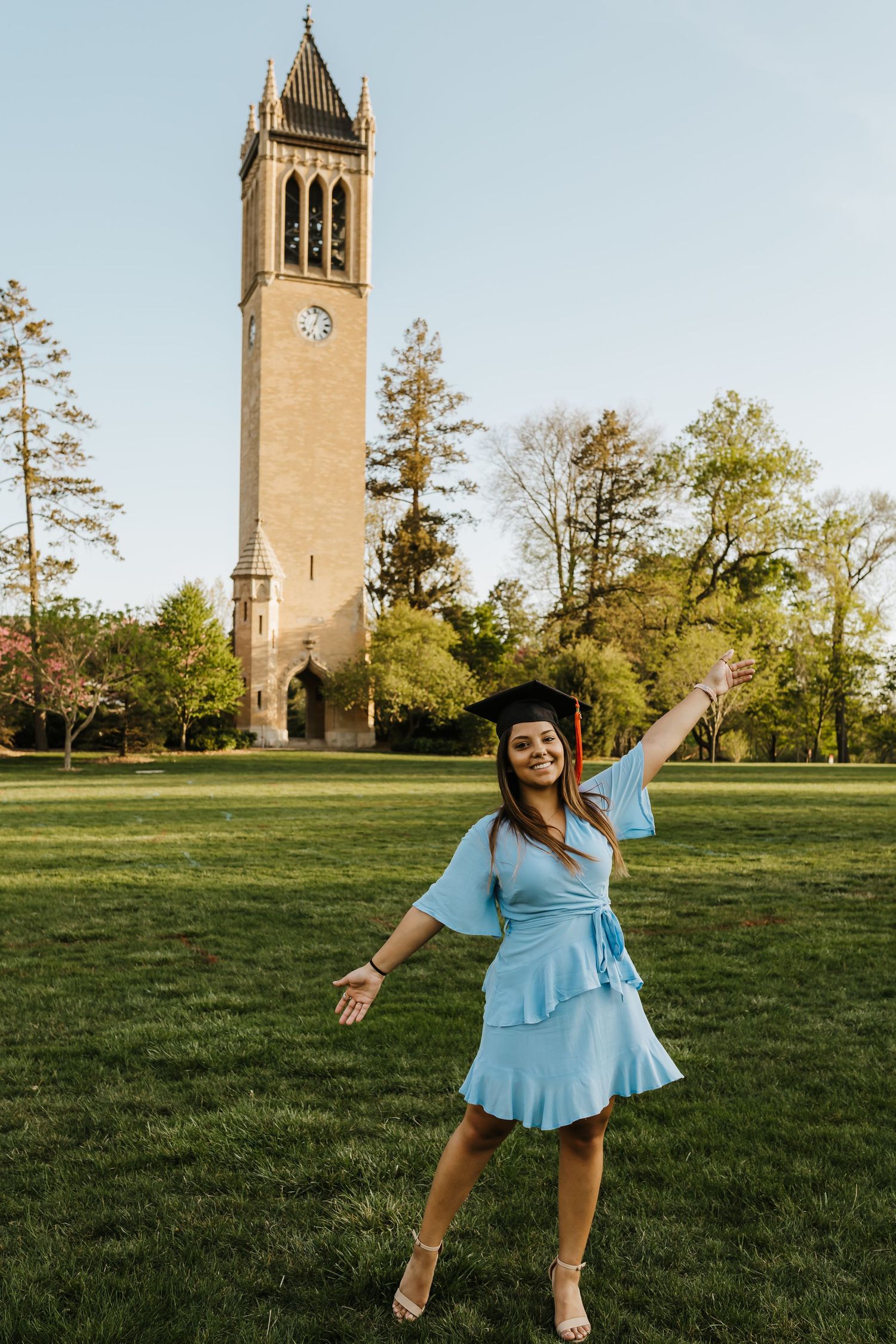 Graduate in light blue dress poses joyfully on lawn with clock tower in background.